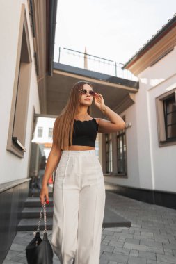 Moda modasına uygun, şık yaz kıyafetleri içinde, beyaz pantolonlu, çantalı, sokakta yürüyen ve güneş gözlüğü takan güzel bir kadın.