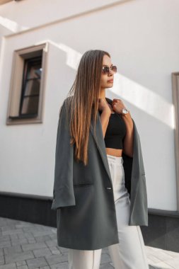 Moda kentsel kıyafetleri içinde güneş gözlüklü zarif genç iş kızı yeşil bir ceket giyiyor ve şehirde beyaz bir duvarın yanında yürüyor. Güzel, şık bir bayan.