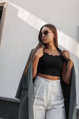 Moda kıyafetleri içinde, güneş gözlüklü, spor ceketli beyaz bir duvarın yanında duran güzel bir kadın manken. Çok zarif bir kız.