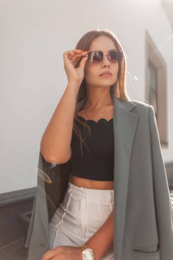 Şık giyinmiş şık, genç bir kadın klasik güneş gözlüklerini takıyor ve güneşli bir günde şehirde oturuyor.