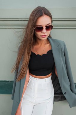 Moda gözlüklü, şık, şık, ceketli genç ve şık bir kadın sokakta yeşil bir duvarın yanında duruyor.