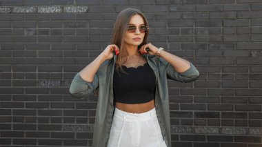 Güneş gözlüklü, yeşil gömlekli, şık, genç bir manken sokakta siyah tuğlalı bir duvarın yanında duruyor.