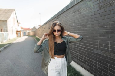 Moda caddesi kıyafetlerinde güneş gözlüğü takan, yeşil gömlekli güzel bir kadın mankeni siyah tuğla bir duvarın yanında yürüyor.