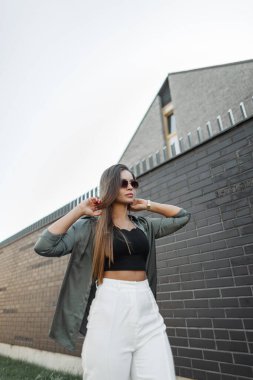 Şık, başarılı, klasik güneş gözlüklü, gömlekli bir iş kadını siyah bir beta duvarın yanında yürüyor ve saçını düzleştiriyor..