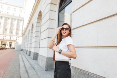 Şık kıyafetler içinde, şık çantalı güzel bir kadın model şehirde yürüyor ve güneş gözlüğü takıyor.