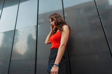 Şık, turuncu tişörtlü, siyah etekli kıvırcık saçlı güzel şehir modeli kız şehirde bir duvarın yanında duruyor. Güzel, şık kadın.