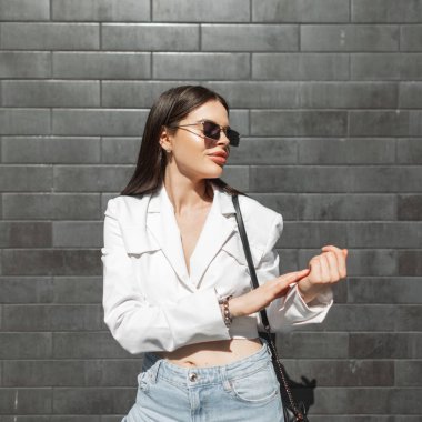 Moda tarzı sokak kıyafetleri içinde geçmişe dönük güneş gözlüklü şık şehir kadını ve güneşli bir günde şehirde modaya uygun çanta taşıyan kot pantolon.