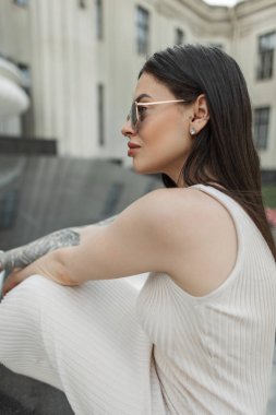 Renkli güneş gözlüklü, bej elbiseli, şık, yeni moda Amerikalı bir kadın dışarıda, sütunları olan bir binanın yanında oturuyor. Güzel kız.