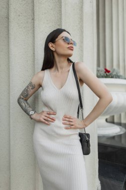 Kolunda bej rengi bir elbise, çanta ve klasik güneş gözlüğü olan güzel moda hippi kadın şehirde bir sütunun yanında duruyor.