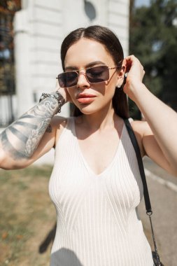 Güneşli bir günde, güzel moda yeniyetme kadın model, modaya uygun giyinmiş, klasik güneş gözlüklü, bej elbiseli, parkta yürüyor.