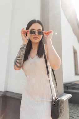 Kolunda bej rengi bir elbise ve çanta olan güzel bir moda hippi kadın şehirdeki bir binanın yakınında klasik güneş gözlüğü takıyor. Yaz güzel kız