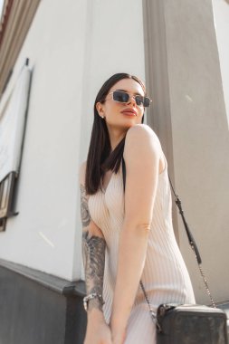 Moda bej elbiseli, modaya uygun güneş gözlüklü, şık, şık, şık bir kadın model sokakta bir duvarın yanında duruyor. Şehirli hippi kız.