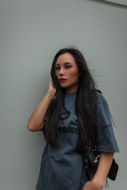 Havalı, havalı, kırmızı dudaklı, şık, şehir kıyafetli, deri çantalı bir Asyalı kadın rüzgarlı bir günde duvarın yanında yürüyor.