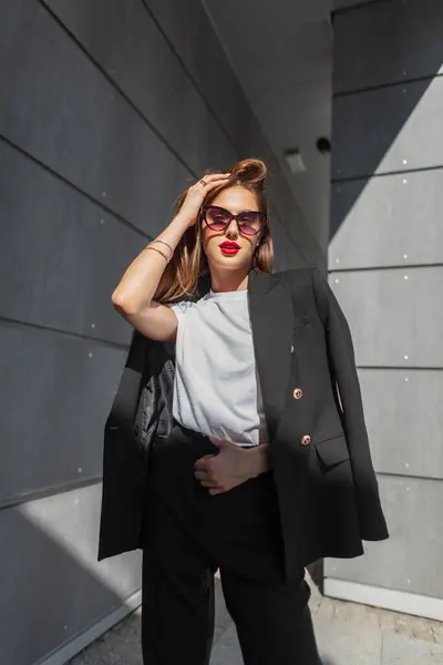 スタイリッシュな美しい若い女性 赤い唇のヴィンテージサングラスとファッションの都市カジュアルな服とTシャツは 日光で灰色の建物の近くに立っています かわいいビジネスガール — ストック写真