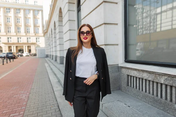 スタイリッシュな黒いブレザーと白のTシャツでサングラスを持つ美しい都市ファッションビジネス女性が通りを歩きます — ストック写真