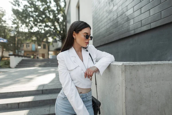 Eski moda güneş gözlüklü, modaya uygun giyinmiş, beyaz ceketli, kot pantolonlu ve şehirde çanta taşıyan güzel bir moda kadınının şehir portresi.