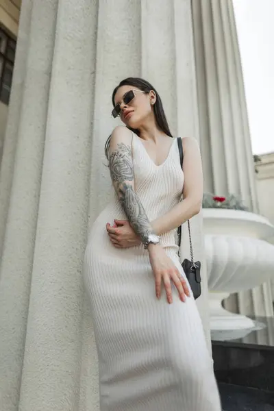 Güneş gözlüklü, bej elbiseli, şık, şık ve şık bir kadın model. Çanta standı ve kentteki bir sütunun yanında poz veriyor. Havalı güzel bayan.