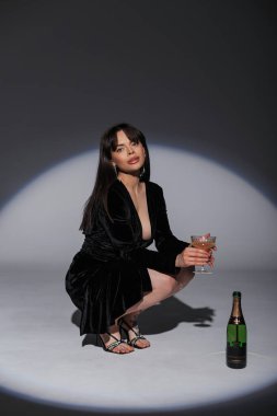 Koyu yakalı siyah elbiseli, göz kamaştırıcı şık şık bir kız stüdyoda bir bardak şampanya ve bir şişe şişeyle oturuyor. Muhteşem güzel bayan kutluyor