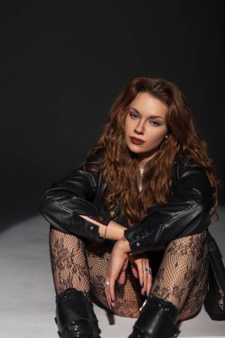 Şık moda seksi kadın siyah deri ceketli, dantel taytlı koyu bir arka planda oturuyor ve poz veriyor.