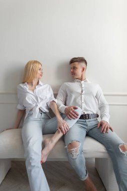 Tarz sahibi genç aşıklar, güzel sarışın bir kız ve yırtık kotlu ve gömlekli şık beyaz elbiseli yakışıklı bir adam evdeki beyaz duvarın yanında oturuyor.
