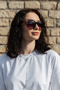 Moda gözlüklü, şık, şık, kıvırcık bir kadın. Beyaz tişörtlü, tuğla bir duvarın yanında.