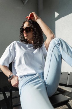 Güneş gözlüklü, beyaz tişörtlü, kot pantolonlu, şık ve genç bir bayan model güneş ışığında şehirde oturuyor ve poz veriyor.