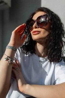 Beyaz tişörtlü şık güneş gözlüklü güzel kıvırcık kız portresi güneş ışığında sokakta oturuyor.