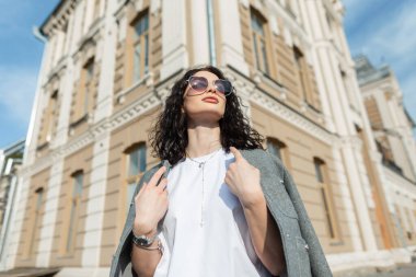 Şık, modaya uygun, kıvırcık kız mankeni, kalp gözlüklü, moda elbiseli, spor ceketli ve binanın yanında beyaz tişörtlü.