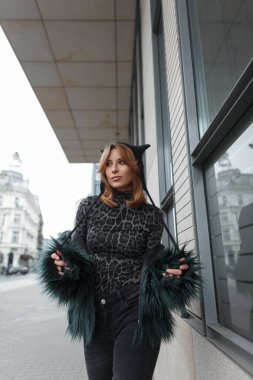 Şık şehir hanımı moda kıyafetleri içinde şapkalı, tüylü ceketli, leopar desenli tişörtlü şehirde yürüyor.