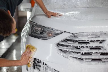 Süngerli bir araba yıkama işçisi araba yıkamada beyaz bir araba yıkar. Araba bakımı ve temizlik