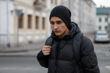 Siyah kışlık giysili, örgü şapkalı, ceketli ve sırt çantalı bir adam şehirde yürüyor.