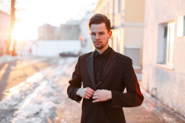 Yakışıklı, başarılı, şık siyah elbiseli genç adam siyah ceketini ilikliyor ve gün batımında karlı bir sokakta duruyor.
