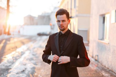 Yakışıklı, başarılı, şık siyah elbiseli genç adam siyah ceketini ilikliyor ve gün batımında karlı bir sokakta duruyor.