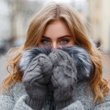 Havalı kış kıyafetleri içinde güzel bir genç kız soğuk bir günde örgü eldivenlerle yüzünü ısıtıyor ve örtüyor.