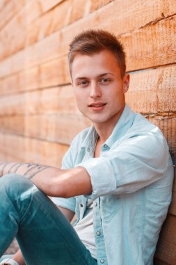 Moda kotlu gömlekli yakışıklı genç adam sokağın ortasında, tahta bir duvarın yanında oturuyor.