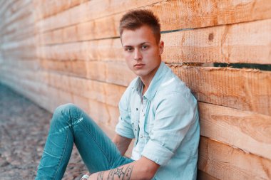Havalı, modaya uygun, modaya uygun kot gömlekli ve kot pantolonlu modaya uygun genç adam ahşap bir arka planın yanında yerde oturuyor.