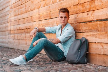 Şık giyinmiş, şık giyinmiş, kotlu, sırt çantalı, spor ayakkabılı yakışıklı bir genç, doğal ahşap duvarın yanında oturuyor.