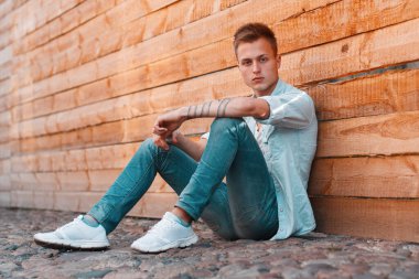 Tişörtlü, kot pantolonlu ve spor ayakkabılı, saç stili olan yakışıklı bir öğrenci ahşap bir duvarın yanında yerde oturuyor.