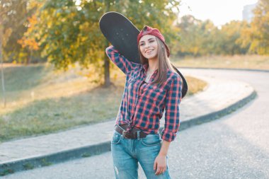 Mutlu, genç, güzel, hippi bir kadın. Şapkalı, moda tişörtlü, kot pantolonlu, elinde kaykayla parkta yürüyen.
