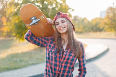 Mutlu genç hippi, şapkalı, moda kıyafetleri içinde, ekose gömlekli, yakışıklı bir kız elinde kaykayla parkta yürüyor.