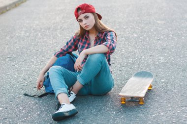 Çizgili tişörtlü, kot pantolonlu, sırt çantalı ve kaykaylı güzel bir Z nesli kadın parkta asfaltta oturuyor.