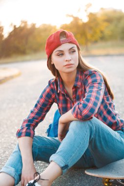Moda sokağı elbiseli, çizgili gömlekli, kaykaylı kot pantolonlu genç ve güzel bir kız gün batımında parktaki asfaltta oturuyor.