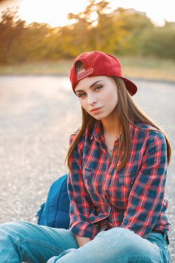 Çizgili gömlekli, sırt çantalı, beysbol şapkalı güzel genç nesil Z kızı gün batımında parktaki asfaltta oturuyor.