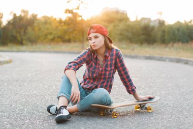 Çizgili gömlekli, kot ve spor ayakkabılı, şapkalı güzel hippi öğrenci Z kuşağı kadın kaykayla asfaltta oturuyor.