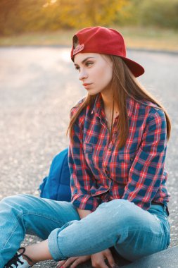 Ekose gömlekli ve sırt çantalı güzel, modaya uygun genç bir bayan öğrenci gün batımında parktaki asfaltta oturuyor.