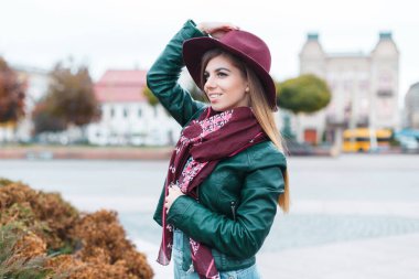 Şık deri ceketli, bordo şapkalı ve klasik bir eşarbı olan neşeli, genç ve güzel bir kız şehirde yürüyor.