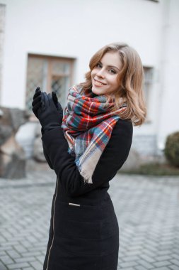 Hoş bir gülümsemesi olan, modaya uygun siyah bir ceketi olan, ekose atkısı ve eldivenleri olan neşeli, güzel bir kız mankeni şehirde geziyor ve eğleniyor.