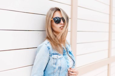 Moda kot ceketli, dantel bluzlu, klasik güneş gözlüklü güzel bir genç kız ahşap bir duvarın yanında duruyor.