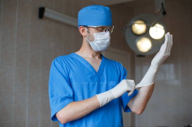 Mavi üniformalı profesyonel doktor eldiven takıp ameliyathanede duruyor.