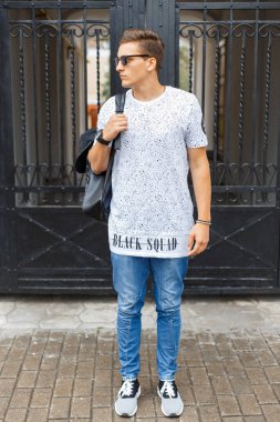 Beyaz tişörtlü, metal bir sokak kapısının yanında sırt çantalı, güneş gözlüklü şık sokak hippisi.